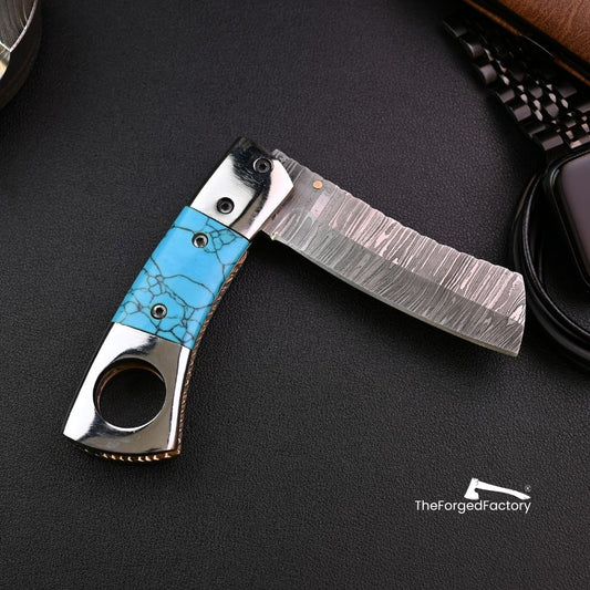 Folding cigar knife with blue handle and silver blade on a dark surface, branded 'The Forged Factory'.