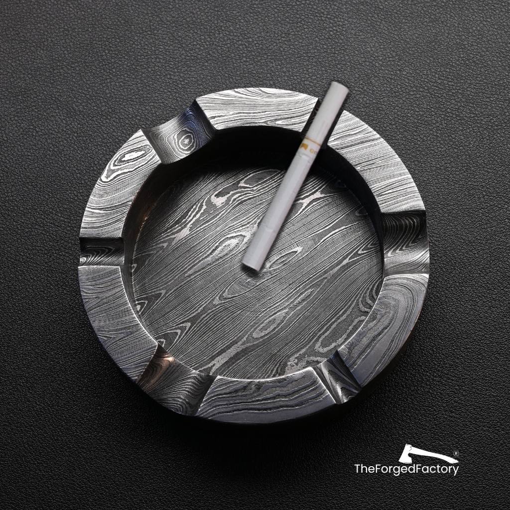 Damascus steel normal ashtray with a cigarette on a black background, branded 'The Forged Factory'.
