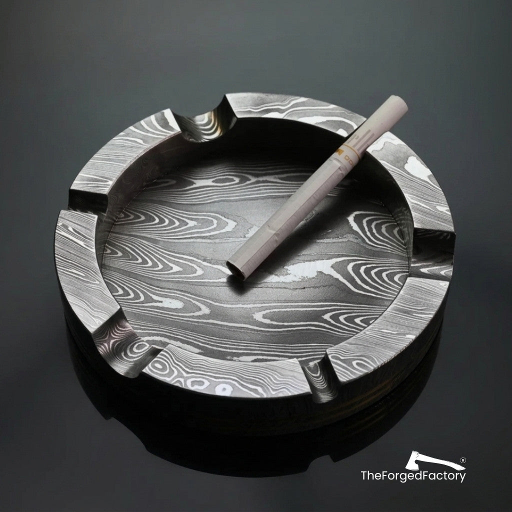 Damascus steel ashtray with a cigarette on a dark background, branded 'The Forged Factory'.