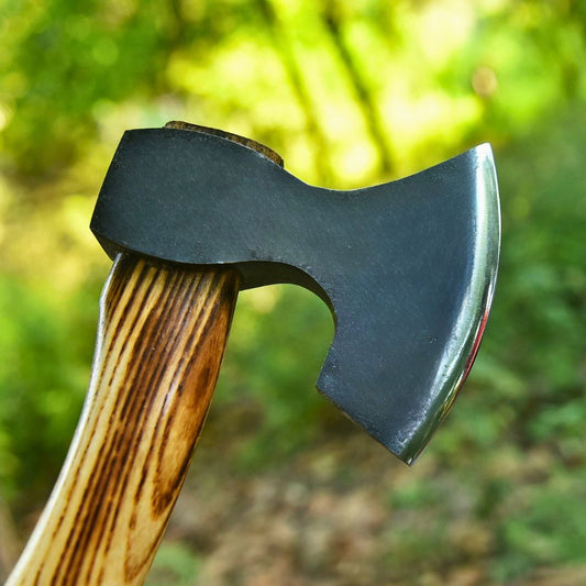A close-up of a hand axe with a dark carbon steel head and a brown wood-grain handle, set against a blurred outdoor forest background.