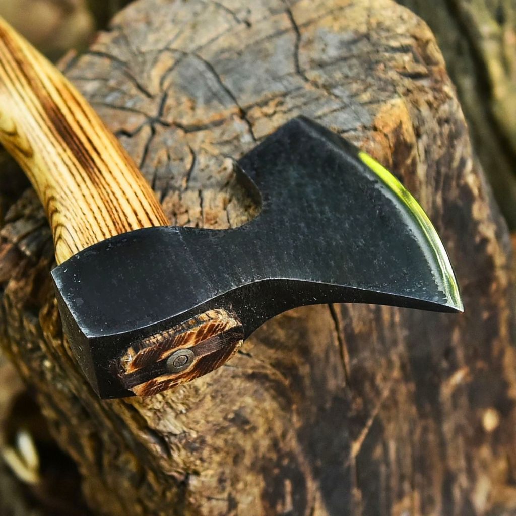 A close-up of a camping bushcraft axe with a black forged steel head and a textured wood handle, resting on a rustic log.