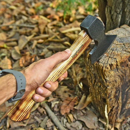 A close-up of a hand holding a brand camping hatchet with a charred wood handle and a black carbon steel head, set against an outdoor background of dry leaves and a tree stump.