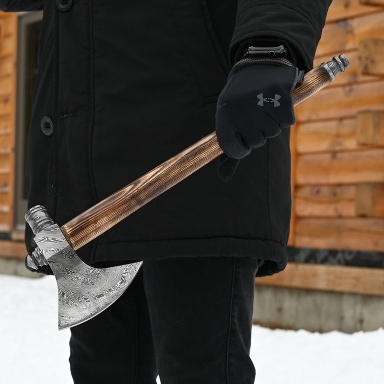 Person holding a hand forged splitting axe with a wooden handle and metal head, wearing a black coat and gloves, against a wooden cabin background.