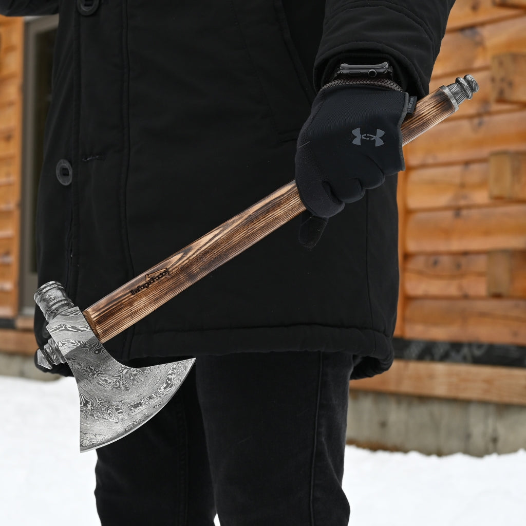 Person holding an axe with a wooden handle in front of a wooden cabin.