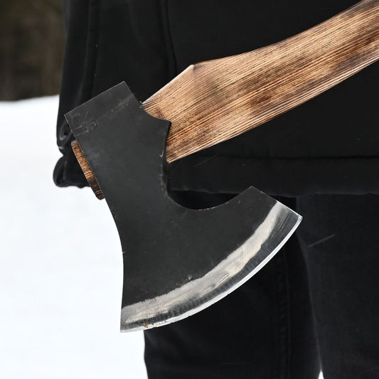 A close-up of a handmade axe with a dark forged steel head and a weathered brown wooden handle, held by a person wearing black clothing against a snowy outdoor background.