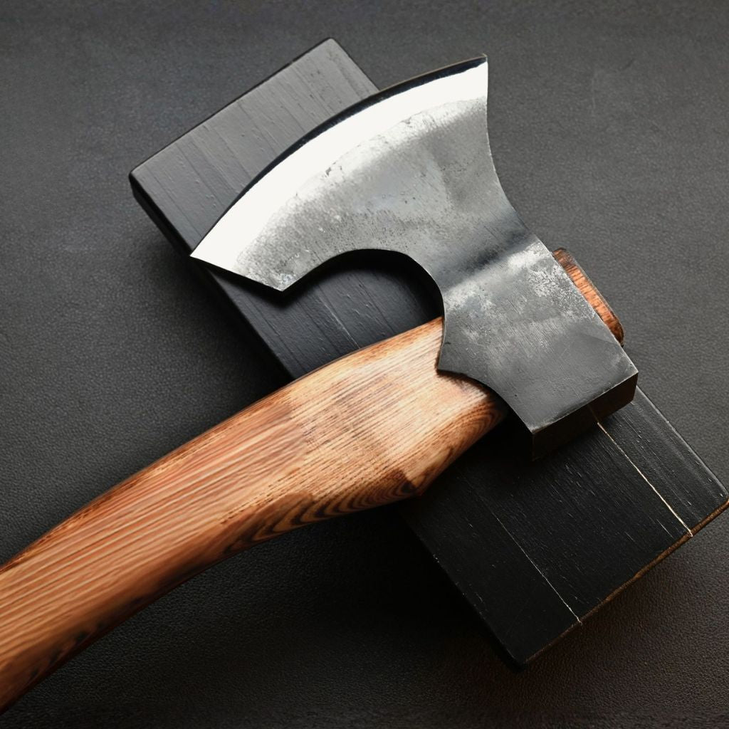 A close-up of a hand forged axe with a polished carbon steel head and a curved wooden handle, resting on a black rectangular block against a dark textured background.