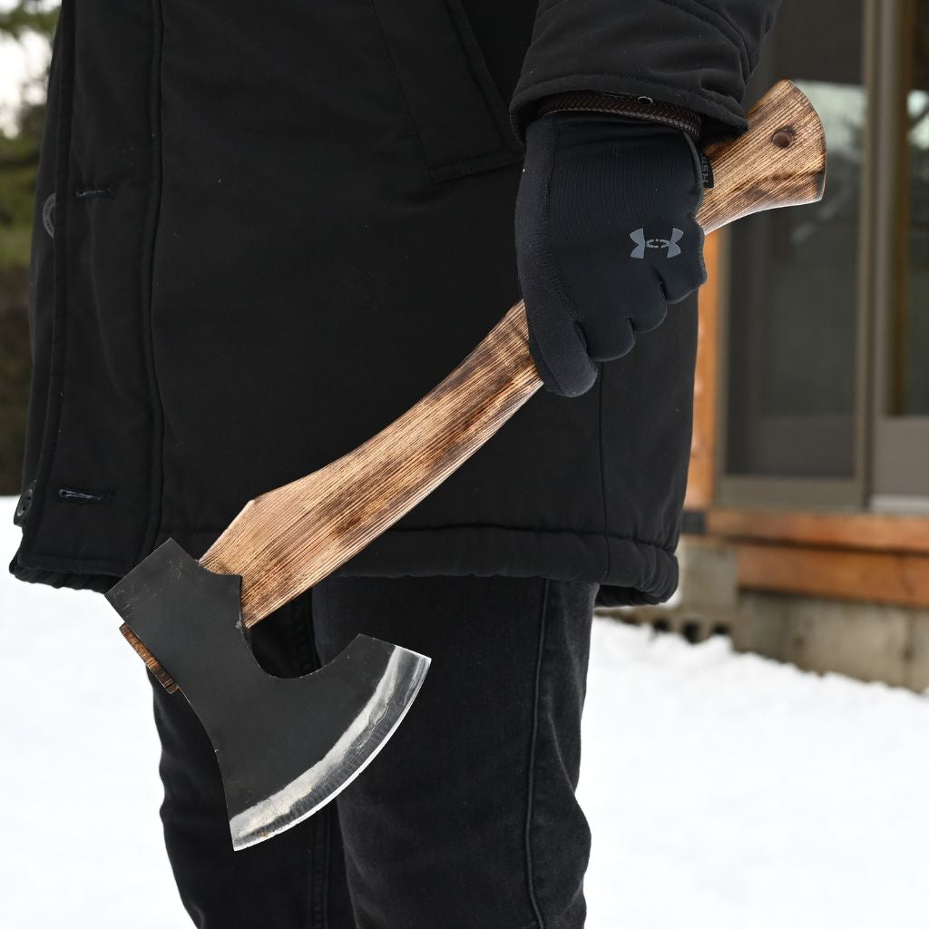 A person in a black winter jacket and black Under Armour gloves holds a hatchet with a dark metal head and a curved brown wooden handle outdoors in snowy weather.