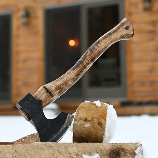 A close-up of a handmade axe with a dark forged carbon steel head and a weathered brown wooden handle, impaled in a log with snow visible, set against a blurred background of a rustic wooden cabin.