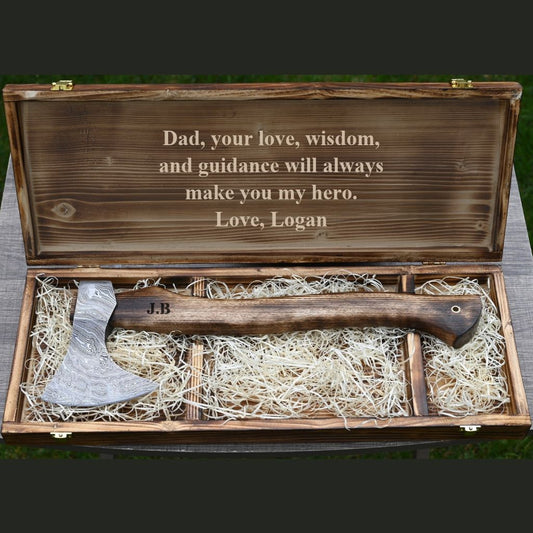 A personalized wooden gift box opened to reveal a small axe resting on shredded paper filler. The inside lid is engraved with a sentimental message for Dad, and the axe handle is personalized with the initials 'J.B'.