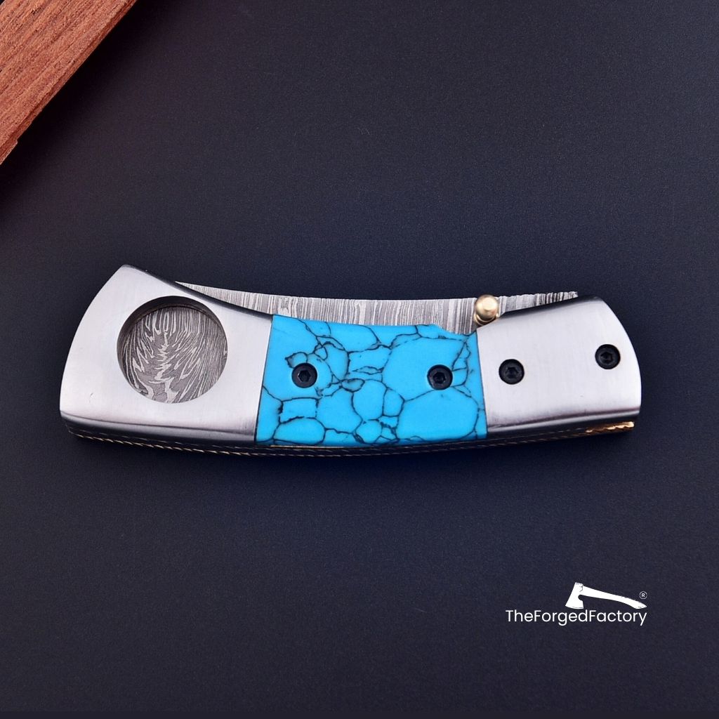 Folding knife with turquoise inlay on a dark surface, branded 'The Forged Factory'.