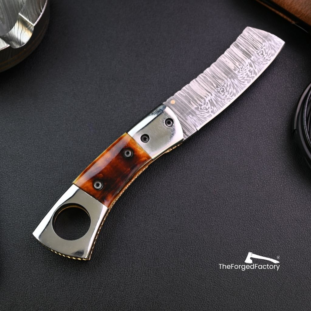 Foldable knife with damascus blade and cigar hole on a dark surface, brand 'The Forged Factory' visible.
